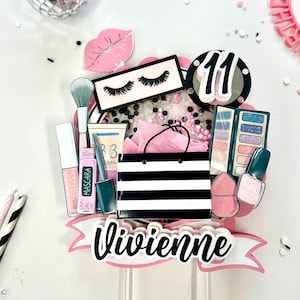 May include: A pink and black cake topper with makeup-themed decorations. The topper includes a black and white striped shopping bag, a makeup palette, lipstick, mascara, and the name "Vivienne". The number "11" is also featured.