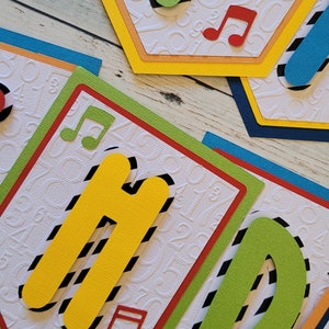 May include: A colorful banner with the word "Happy" spelled out in large, cut-out letters. Each letter is decorated with black and white stripes and a musical note. The banner is made of cardstock and has a white background with a subtle embossed pattern of numbers.