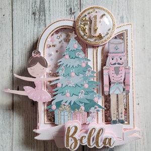 May include: A layered papercraft design featuring a ballerina, Christmas tree, and nutcracker. The design includes the name "Bella" in gold glitter lettering, a number "1" in a clear circle, and pink and gold accents. The background is a light wood grain.
