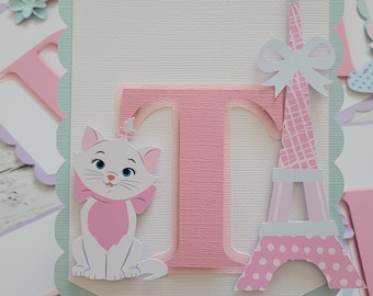 May include: A pink and white banner with a scalloped edge. The banner features a pink letter T, a white cat with a pink bow, and a pink and white Eiffel Tower. The banner is perfect for a birthday party.