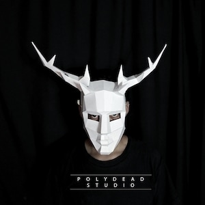 May include: A white paper mask with antlers, resembling a deer, with a geometric design. The mask is worn by a person with a black shirt and the text "POLYDEAD STUDIO" is visible below the mask.