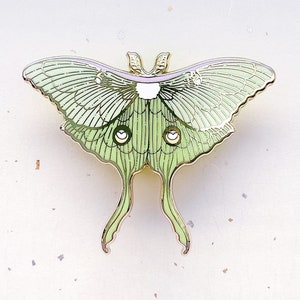 Puede incluir: Un pin de metal dorado con una polilla luna verde y blanca con alas y antenas detalladas.