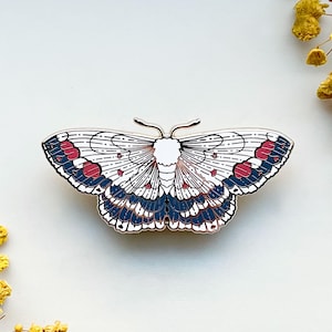 May include: A detailed enamel pin in the shape of a moth. The pin features a white body and wings with intricate designs in navy blue, red, and black. The pin is a decorative accessory.