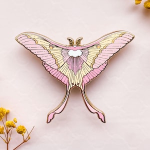 May include: A gold-colored moth pin with pink and yellow wings. The pin features intricate details and a delicate design. The moth's wings are spread wide, showcasing the detailed patterns. The pin is a decorative accessory.