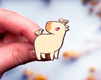 May include: Gold enamel pin featuring a cartoon capybara with three small yellow birds perched on its head. The capybara is light brown with a pink nose and black eyes.