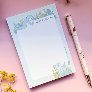 May include: A light blue notepad with the text "Stuff I Otter Do" and an illustration of an otter. A white pen with a gold clip and a pattern of foxes, stars, and fish is next to the notepad. The background is pink.