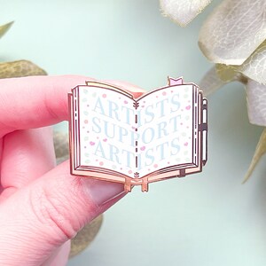 May include: A rose gold enamel pin shaped like an open book with the words "Artists Support Artists" on the pages. The pin has a white background with pink and blue floral accents.