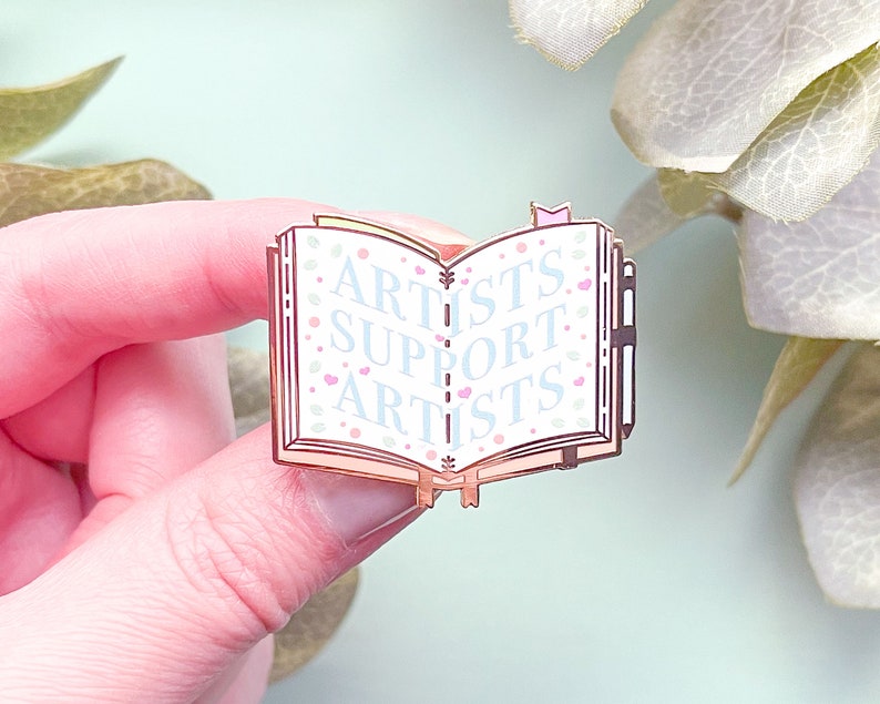 May include: A rose gold enamel pin shaped like an open book with the words "Artists Support Artists" on the pages. The pin has a white background with pink and blue floral accents.