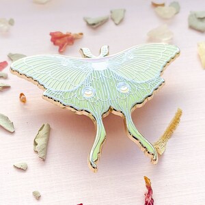 Puede incluir: Un pin de esmalte dorado que representa una polilla luna verde con detalles blancos. La polilla se representa con las alas extendidas, mostrando detalles intrincados y un acabado delicado e iridiscente.