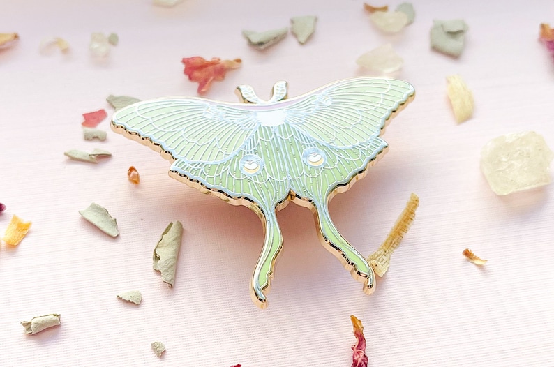Puede incluir: Un pin de esmalte dorado que representa una polilla luna verde con detalles blancos. La polilla se representa con las alas extendidas, mostrando detalles intrincados y un acabado delicado e iridiscente.