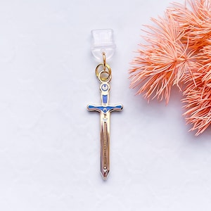 May include: A gold-coloured sword charm with blue enamel accents and a clear plastic clasp. The sword has a detailed hilt and blade, with a small gemstone near the centre. The charm is suspended from a gold-coloured ring.