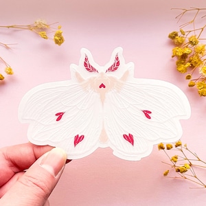 May include: White moth sticker with pink hearts on its wings. The moth has a delicate, intricate design and is perfect for adding a touch of whimsy to any project.