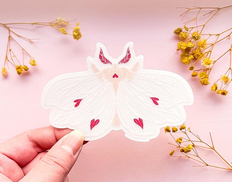 May include: White moth sticker with pink hearts on its wings. The moth has a delicate, intricate design and is perfect for adding a touch of whimsy to any project.