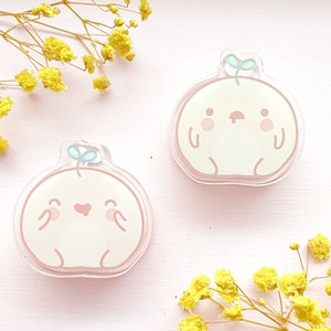 May include: Two peach-colored cartoon characters with happy and sad expressions. Each character has a small green sprout on top of their head. The characters are made of clear acrylic and are on a pink background.