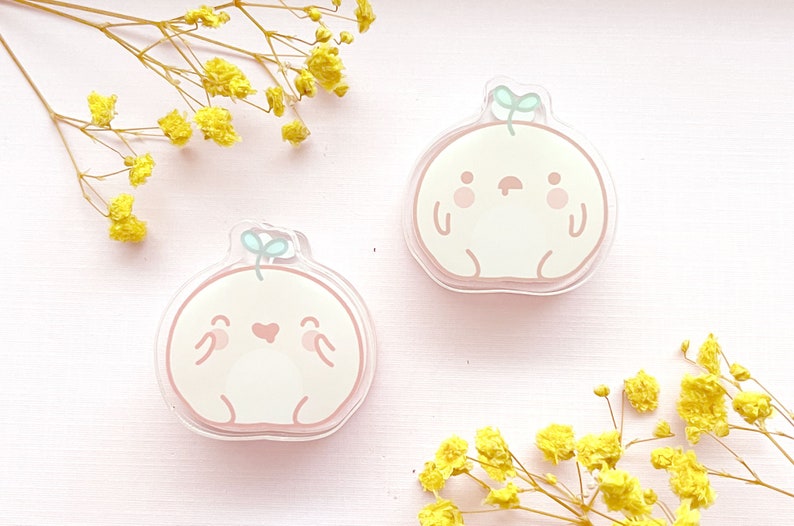 May include: Two peach-colored cartoon characters with happy and sad expressions. Each character has a small green sprout on top of their head. The characters are made of clear acrylic and are on a pink background.