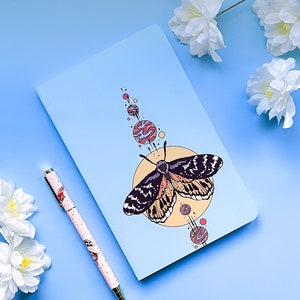 May include: A blue notebook with a colorful butterfly illustration and a pen with a floral pattern. The butterfly is surrounded by a yellow circle with smaller circles around it. The notebook is on a light blue background.