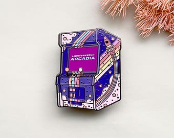 May include: Enamel pin featuring a retro arcade machine design. The pin is primarily purple with a pink and rainbow color scheme, and the text "LIGHTSPEED ARCADIA" is displayed on the screen. A rocket ship is also featured.