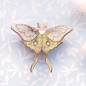 Puede incluir: Un pin de metal de color dorado con forma de polilla luna con un degradado rosa y amarillo. La polilla tiene alas y antenas detalladas.