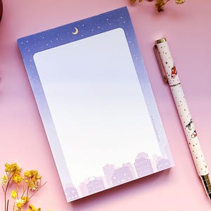 May include: A purple notepad with a cityscape and a crescent moon with stars on the top. The notepad is on a pink surface with a gold pen with a fish design.