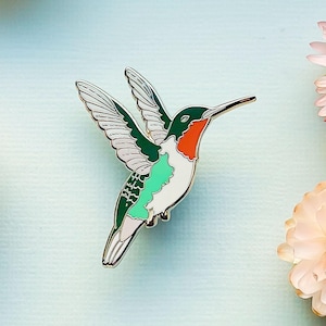 May include: A green, white, and red enamel hummingbird pin with a long, thin beak. The hummingbird is in flight with its wings spread out.