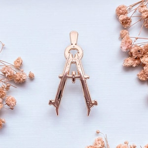 Puede incluir: Un alfiler de compás de color oro rosa, una herramienta de dibujo, está centrado en un fondo azul claro. El alfiler está rodeado de flores secas de color marrón claro. El compás tiene dos patas conectadas en la parte superior con un asa.