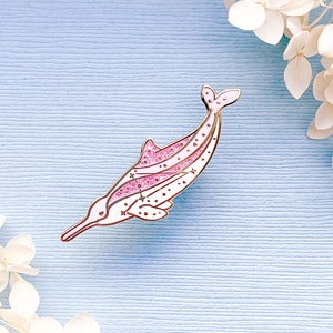 Op de afbeelding: Een roze en witte emaille pin in de vorm van een rivierdolfijn. De pin heeft een gouden omtrek, roze glitteraccenten en kleine sterren. De dolfijn staat tegen een lichtblauwe achtergrond met witte bloemen.