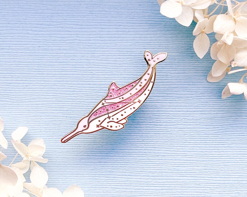 Op de afbeelding: Een roze en witte emaille pin in de vorm van een rivierdolfijn. De pin heeft een gouden omtrek, roze glitteraccenten en kleine sterren. De dolfijn staat tegen een lichtblauwe achtergrond met witte bloemen.