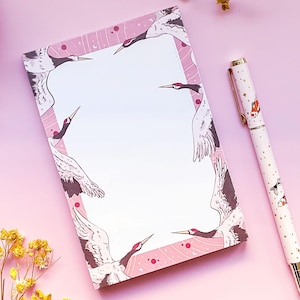 May include: Pink notepad with a white interior and a border of flying cranes. The cranes are in shades of brown and gray against a pink background with white stars and dots.