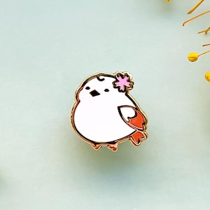 May include: A gold-colored metal pin in the shape of a cartoon bird. The bird is white with orange and pink accents, a black beak, and a pink flower on its head. The pin is on a light blue background.