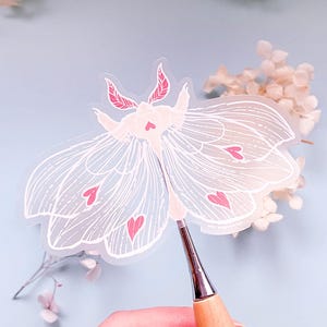 May include: A clear vinyl sticker of a white moth with pink hearts. The moth has delicate wings and a detailed design.