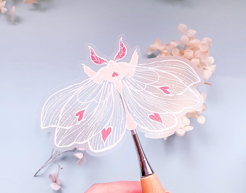 May include: A clear vinyl sticker of a white moth with pink hearts. The moth has delicate wings and a detailed design.