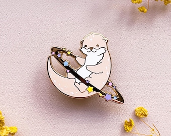 May include: An enamel pin depicting a light brown otter on a crescent moon. The otter holds a star-studded wand, with colourful stars on the moon's ring. The pin has a gold outline and is set against a soft pink background. A cute, celestial-themed accessory.