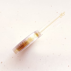 Puede incluir: Un rollo de cinta washi transparente con un patrón decorativo dorado. La cinta está sobre un fondo blanco.