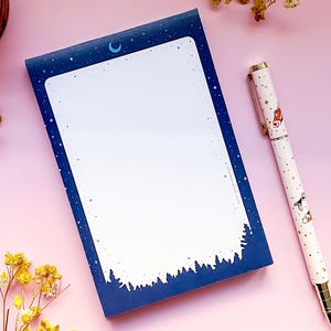 May include: A blue notepad with a crescent moon and stars design. The notepad has a white lined interior and is perfect for writing down notes or ideas. A pen with a white background and a pattern of goldfish is lying on a pink surface next to the notepad.