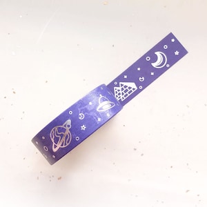 May include: A roll of purple washi tape with silver celestial and architectural designs. The tape features illustrations of planets, stars, pyramids, a bell, and a crescent moon. The tape is unrolled on a white surface.