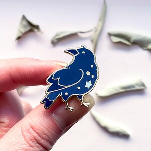 May include: A dark blue enamel pin in the shape of a crow, with a gold outline and white star details. The pin is held between fingers against a light pink background, with dried leaves in the background.