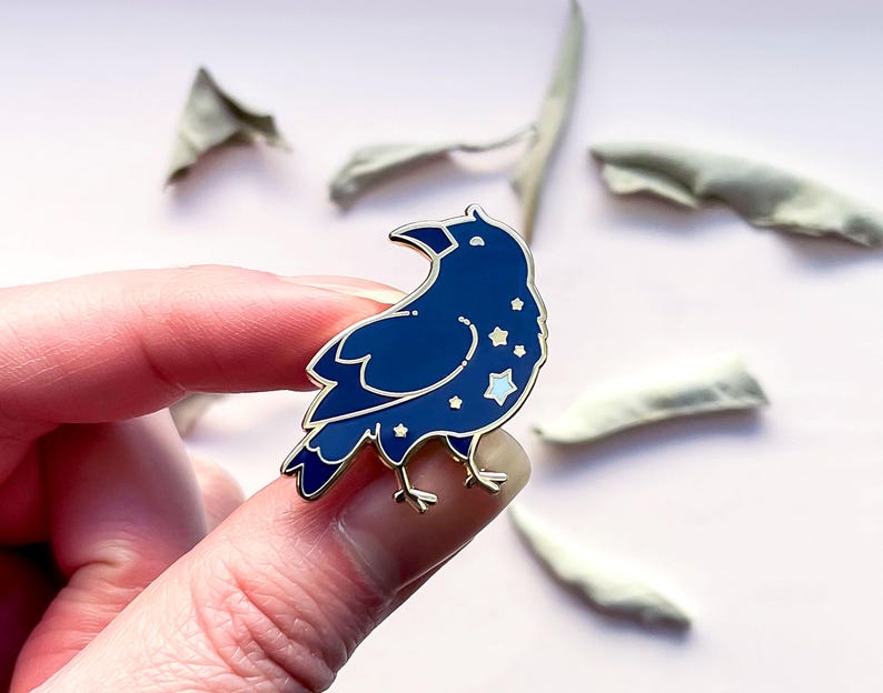 May include: A dark blue enamel pin in the shape of a crow, with a gold outline and white star details. The pin is held between fingers against a light pink background, with dried leaves in the background.
