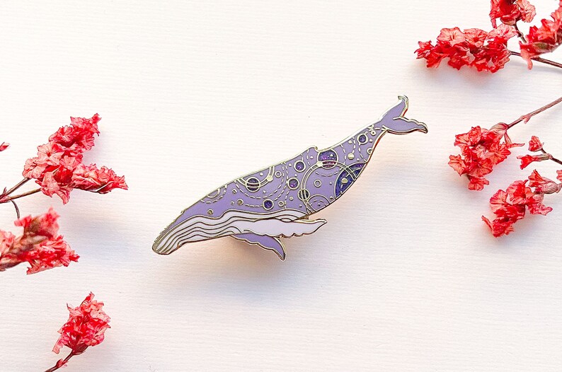 May include: A purple and gold enamel pin of a whale with a galaxy design. The whale is swimming to the right and has a detailed design of stars, planets, and constellations.