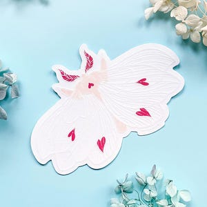 May include: White paper sticker of a moth with pink hearts on its wings. The moth has a detailed design and is a whimsical illustration.