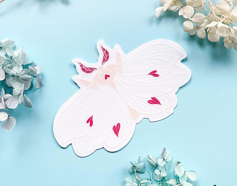 May include: White paper sticker of a moth with pink hearts on its wings. The moth has a detailed design and is a whimsical illustration.