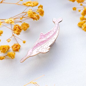 Op de afbeelding: Een roze en witte emaille pin in de vorm van een dolfijn. De pin heeft een gouden omtrek en glitteraccenten. De dolfijn zwemt naar links. De achtergrond is lichtblauw met gele bloemen. De pin is ongeveer 5 cm lang.