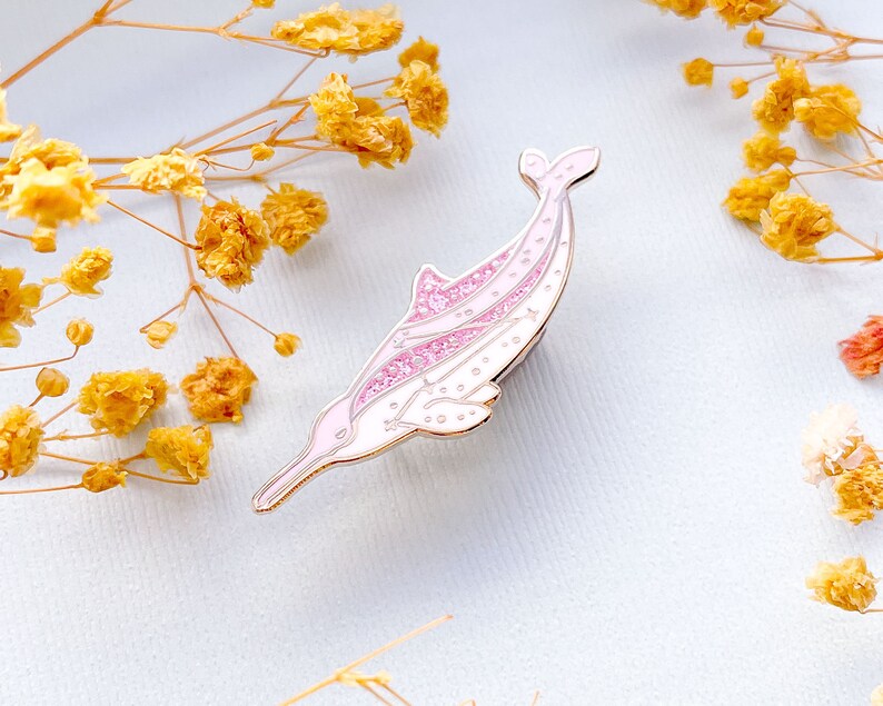 Op de afbeelding: Een roze en witte emaille pin in de vorm van een dolfijn. De pin heeft een gouden omtrek en glitteraccenten. De dolfijn zwemt naar links. De achtergrond is lichtblauw met gele bloemen. De pin is ongeveer 5 cm lang.
