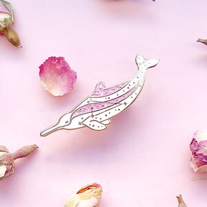 Op de afbeelding: Een witte en roze emaille pin in de vorm van een dolfijn. De dolfijn heeft een lange snuit en een sterrenontwerp op zijn lichaam. De pin is omgeven door gedroogde rozenknoppen en bloemblaadjes op een roze achtergrond.