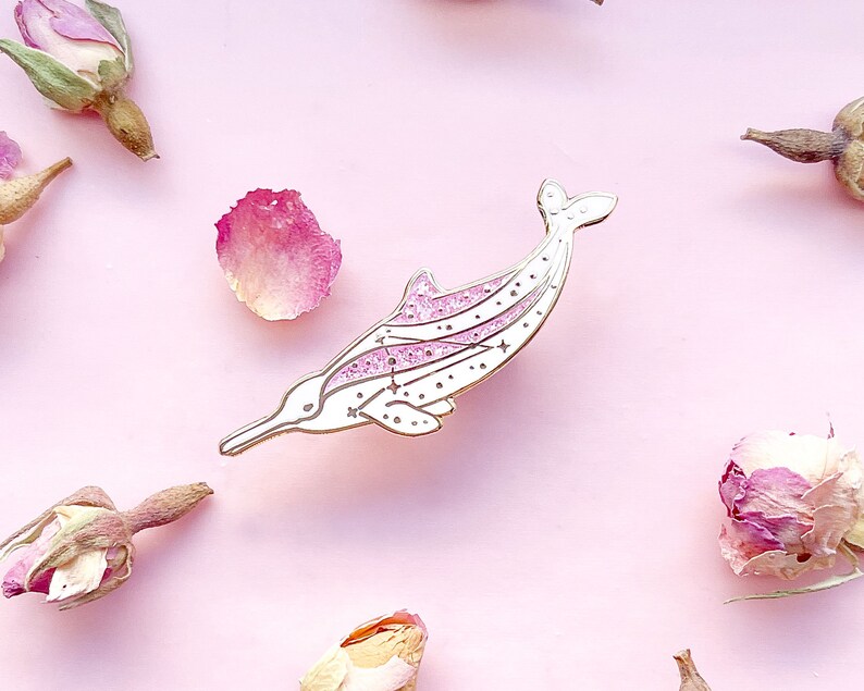 Op de afbeelding: Een witte en roze emaille pin in de vorm van een dolfijn. De dolfijn heeft een lange snuit en een sterrenontwerp op zijn lichaam. De pin is omgeven door gedroogde rozenknoppen en bloemblaadjes op een roze achtergrond.