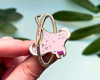 May include: A gold-colored enamel pin featuring a pink manta ray design with star and dot details. The manta ray is set within a gold frame. The pin has a shiny, polished finish.