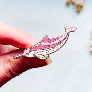 Op de afbeelding: Een roze en gouden dolfijn emaille pin. De pin heeft een gestileerd dolfijnontwerp met glitteraccenten en sterdetails. De dolfijn is lichtroze met donkerdere roze accenten en gouden contouren.