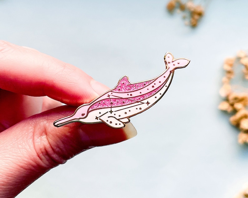 Op de afbeelding: Een roze en gouden dolfijn emaille pin. De pin heeft een gestileerd dolfijnontwerp met glitteraccenten en sterdetails. De dolfijn is lichtroze met donkerdere roze accenten en gouden contouren.