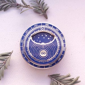Peut inclure: Une épingle en métal doré avec un design en émail bleu. L'épingle présente un design circulaire avec un calendrier autour du bord extérieur. Le centre de l'épingle présente une représentation stylisée de la lune et des étoiles. L'épingle présente également le mot "infini" au centre.
