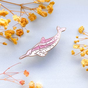 Op de afbeelding: Een roze en witte dolfijn emaille pin met glitter accenten en ster details. De pin heeft een gouden omtrek. Geplaatst tegen een lichtblauwe achtergrond met gedroogde gele bloemen.