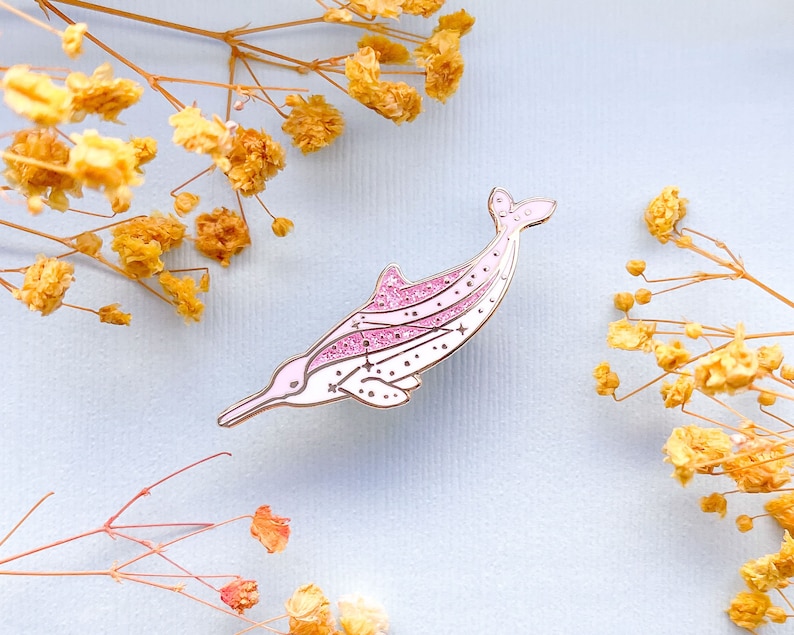 Op de afbeelding: Een roze en witte dolfijn emaille pin met glitter accenten en ster details. De pin heeft een gouden omtrek. Geplaatst tegen een lichtblauwe achtergrond met gedroogde gele bloemen.